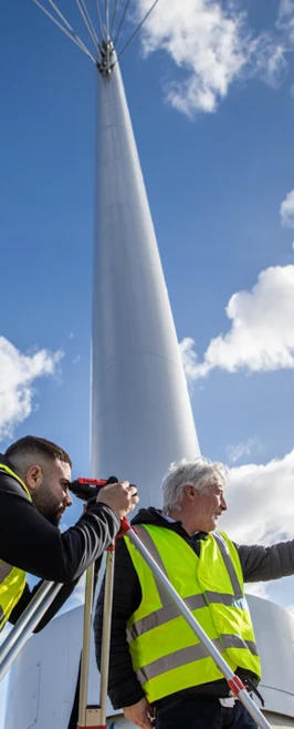 A student using a theodolite for a land surveying exercise, with a lecturer pointing towards an area of interest. Another student holds a measuring staff, all wearing high-visibility vests on a modern bridge. A student using a theodolite for a land surveying exercise, with a lecturer pointing towards an area of interest. Another student holds a measuring staff, all wearing high-visibility vests on a modern bridge.